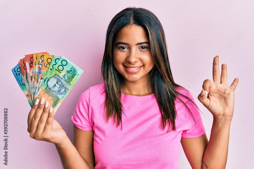 Young latin girl holding australian dollars doing ok sign with fingers, smiling friendly gesturing excellent symbol Young latin girl holding australian dollars doing ok sign with fingers, smiling friendly gesturing excellent symbol