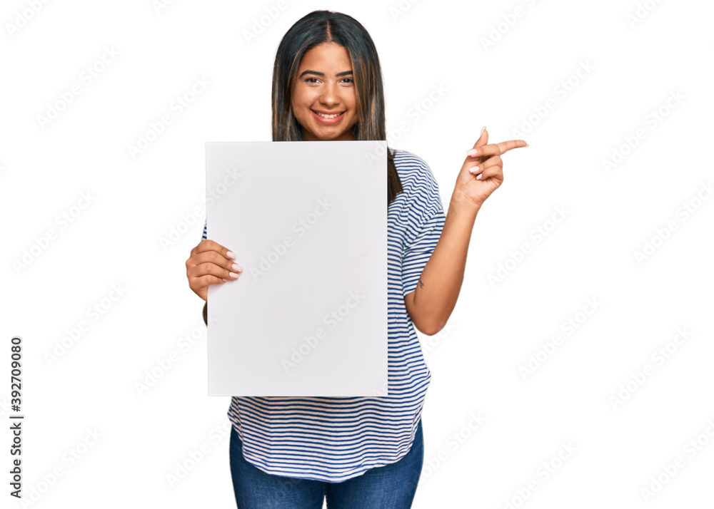 Young latin girl holding blank empty banner smiling happy pointing with hand and finger to the side Young latin girl holding blank empty banner smiling happy pointing with hand and finger to the side