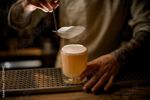 barman holds tweezers with decorative leaf and accurate decorate glass of alcoholic cocktail