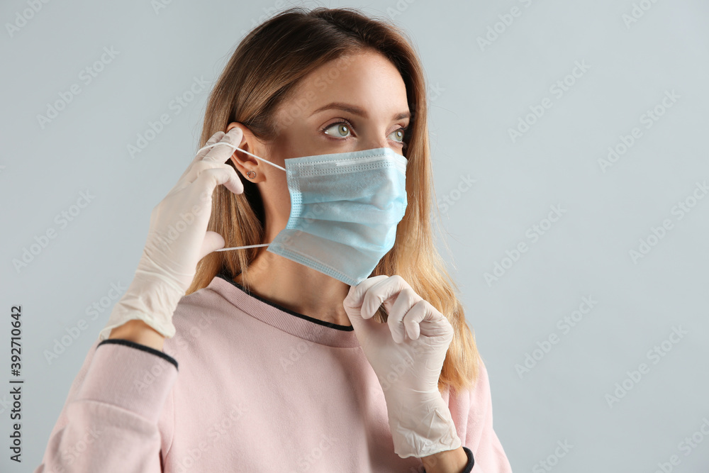 Young woman in medical gloves putting on protective mask against grey background