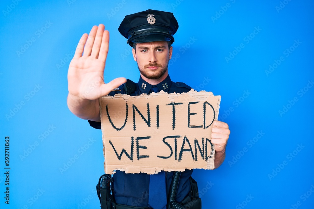 Young caucasian man wearing police uniform holding united we stand banner with open hand doing stop sign with serious and confident expression, defense gesture Young caucasian man wearing police uniform holding united we stand banner with open hand doing stop sign with serious and confident expression, defense gesture