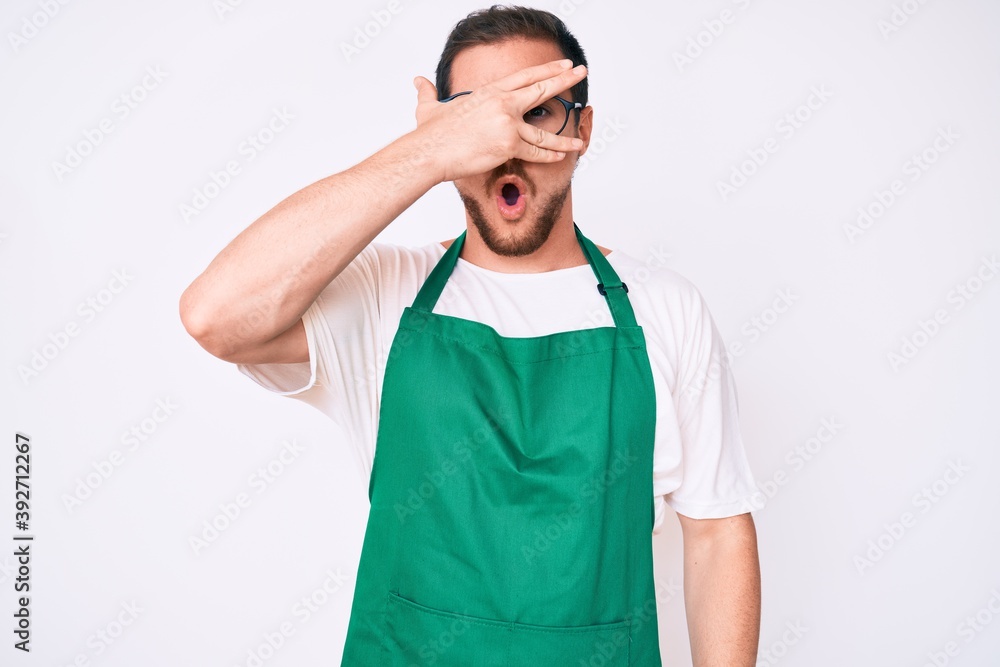 Fototapeta premium Young handsome man wearing apron peeking in shock covering face and eyes with hand, looking through fingers afraid