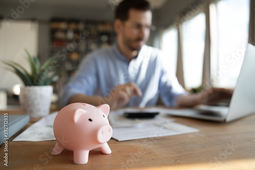 Man sit at desk manage expenses, calculate expenditures, pay bills online use laptop, makes household finances analysis, close up focus on pink piggy bank. Save money for future, be provident concept