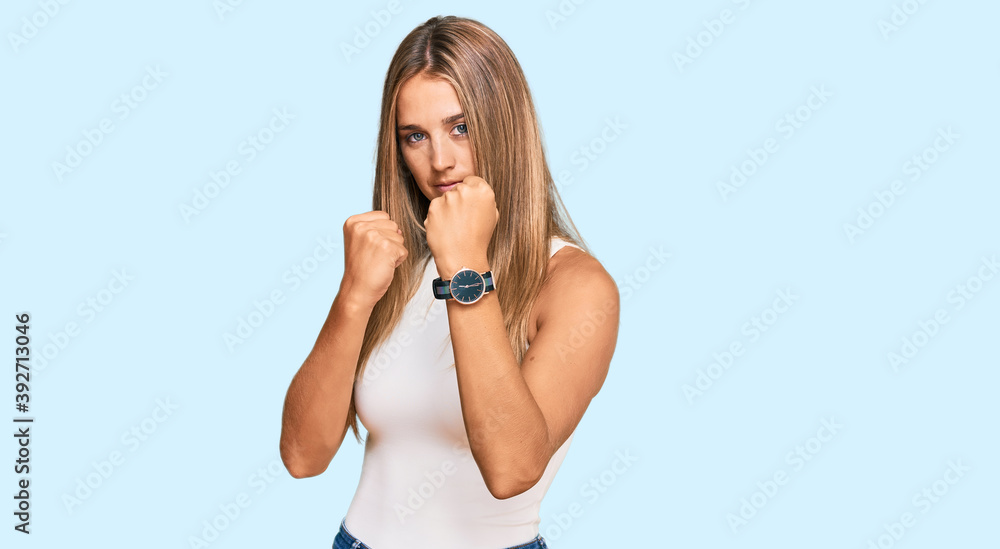 Young blonde woman wearing casual style with sleeveless shirt ready to fight with fist defense gesture, angry and upset face, afraid of problem Young blonde woman wearing casual style with sleeveless shirt ready to fight with fist defense gesture, angry and upset face, afraid of problem