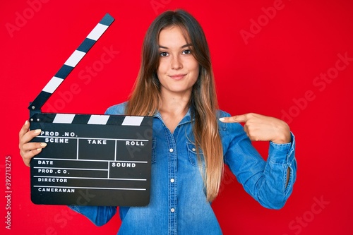 Beautiful caucasian woman holding video film clapboard pointing finger to one self smiling happy and proud