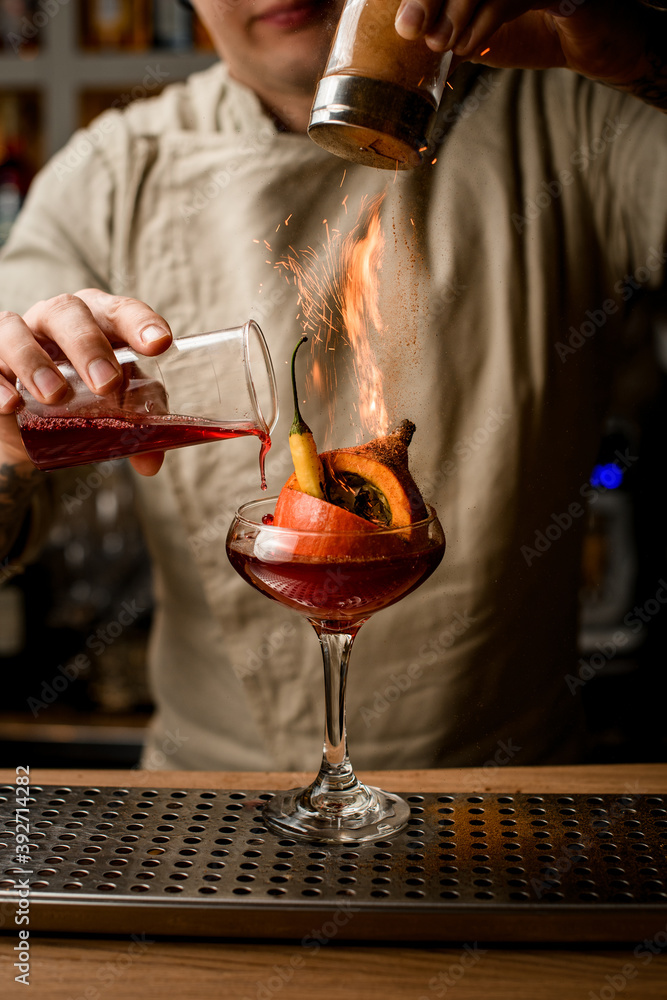 man bartender add ingredients to glass with pumpkin and pepper inside and makes fire over it