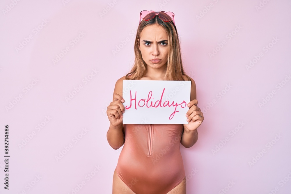 Young beautiful blonde woman wearing swimwear and holding holidays banner skeptic and nervous, frowning upset because of problem. negative person. Young beautiful blonde woman wearing swimwear and holding holidays banner skeptic and nervous, frowning upset because of problem. negative person.