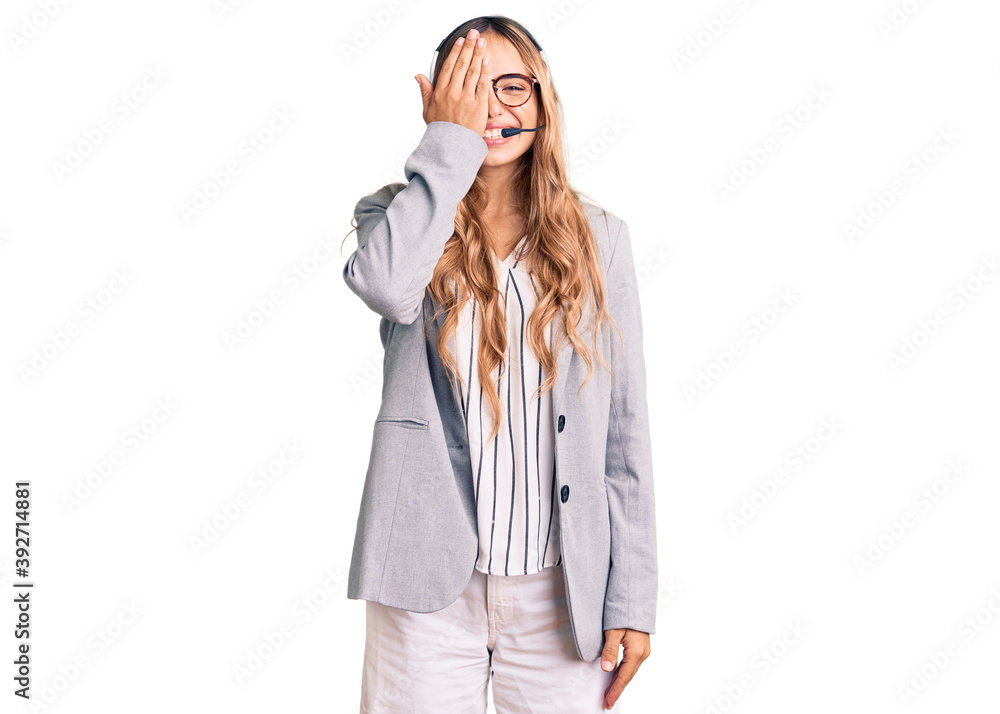 Young beautiful blonde woman wearing call center agent headset covering one eye with hand, confident smile on face and surprise emotion. Young beautiful blonde woman wearing call center agent headset covering one eye with hand, confident smile on face and surprise emotion.