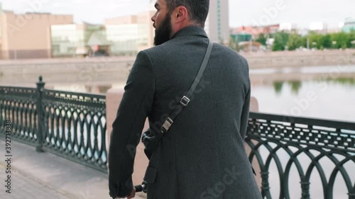 Tilting up and down footage of adult man dressed in smart clothes riding electric scooter fast down bridge in city centre with river and modern museum building in background