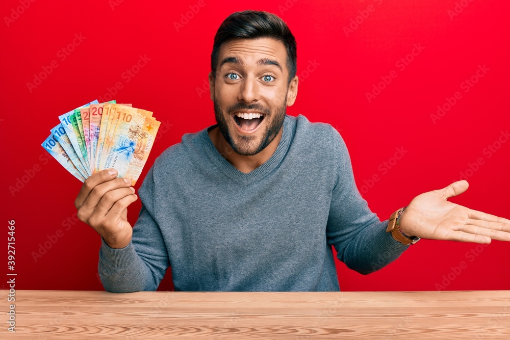 Handsome hispanic man holding swiss franc banknotes celebrating achievement with happy smile and winner expression with raised hand Handsome hispanic man holding swiss franc banknotes celebrating achievement with happy smile and winner expression with raised hand