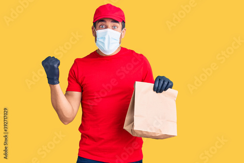 Young hispanic man delivering food wearing covid-19 safety mask holding paper bag screaming proud, celebrating victory and success very excited with raised arms