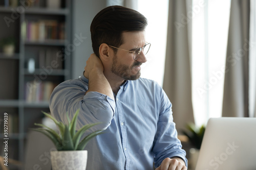 Young man sit at workplace desk touch neck feeling ache, massaging nape to relief painful feelings. Poor sitting posture during laptop usage, sedentary lifestyle, chronic disorder fibromyalgia concept
