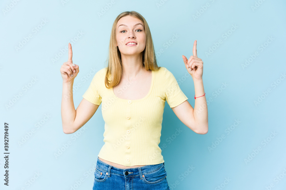 Young blonde woman isolated on blue background indicates with both fore fingers up showing a blank space. Young blonde woman isolated on blue background indicates with both fore fingers up showing a blank space.