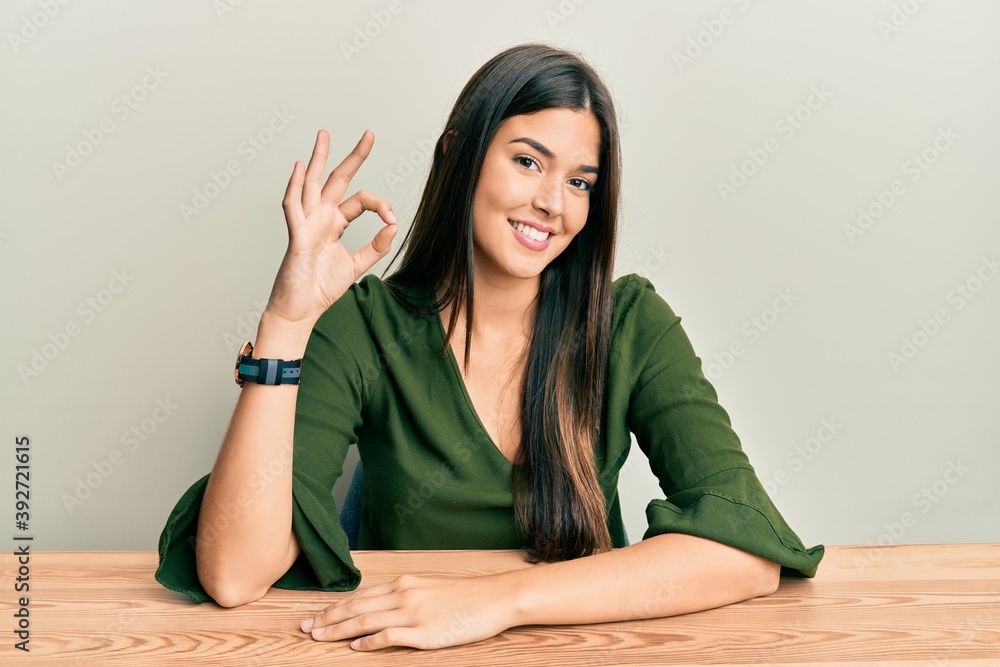 Young brunette woman wearing casual clothes sitting on the table smiling positive doing ok sign with hand and fingers. successful expression. Young brunette woman wearing casual clothes sitting on the table smiling positive doing ok sign with hand and fingers. successful expression.