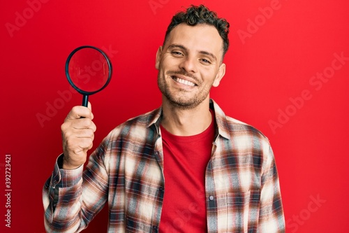 Handsome man with beard holding magnifying glass looking positive and happy standing and smiling with a confident smile showing teeth © Krakenimages.com