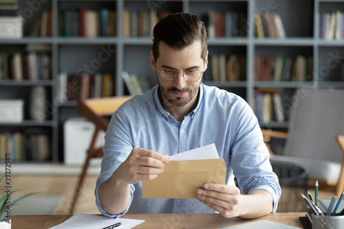 Cuadro en lienzo Serious businessman sit at workplace office desk holding envelope take out letter feels interested read business news, got invitation, learns bank statement information
