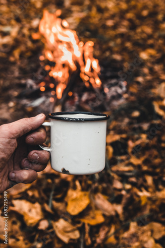 Man hand holds a hot cup of coffee at the background of the campfire. Concept adventure active vacations outdoor. Summer camp.