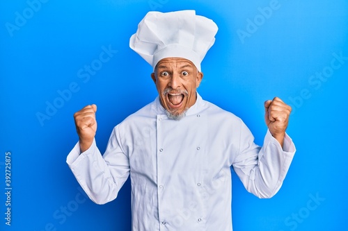 Middle age grey-haired man wearing professional cook uniform and hat celebrating surprised and amazed for success with arms raised and open eyes. winner concept.