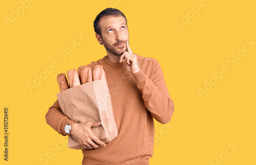 Young handsome man holding paper bag with bread serious face thinking about question with hand on chin, thoughtful about confusing idea