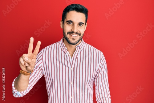 Young hispanic man wearing business shirt smiling with happy face winking at the camera doing victory sign. number two.