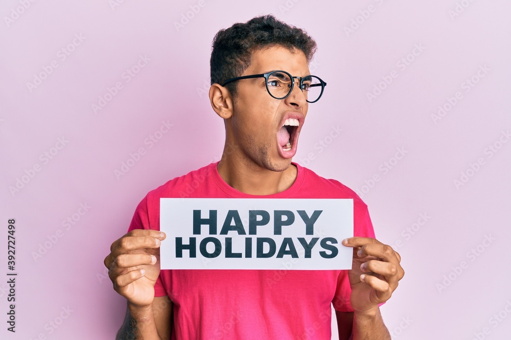 Young handsome african american man holding happy holidays message angry and mad screaming frustrated and furious, shouting with anger. rage and aggressive concept. Young handsome african american man holding happy holidays message angry and mad screaming frustrated and furious, shouting with anger. rage and aggressive concept.