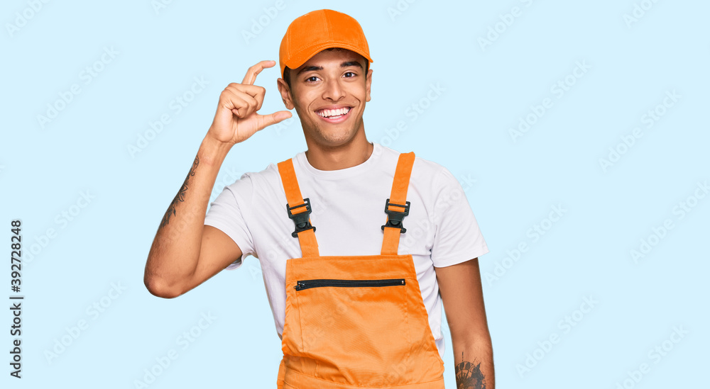 Young handsome african american man wearing handyman uniform smiling and confident gesturing with hand doing small size sign with fingers looking and the camera. measure concept. Young handsome african american man wearing handyman uniform smiling and confident gesturing with hand doing small size sign with fingers looking and the camera. measure concept.