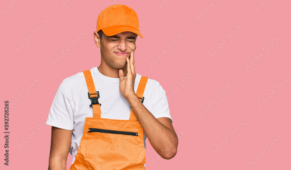 Young handsome african american man wearing handyman uniform touching mouth with hand with painful expression because of toothache or dental illness on teeth. dentist