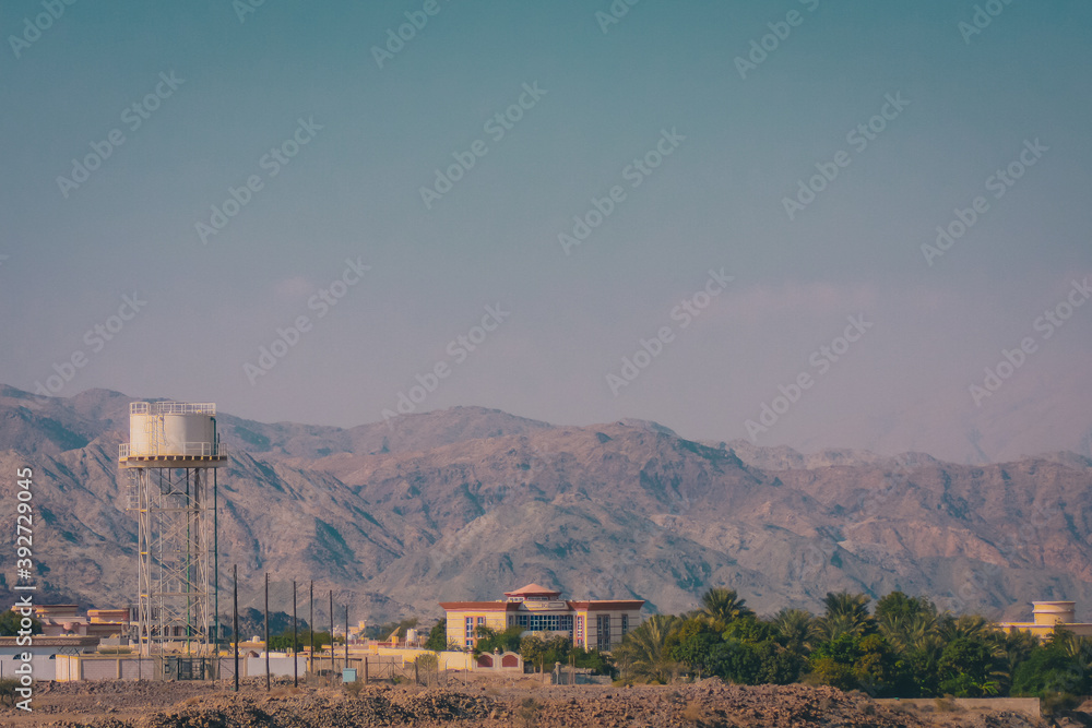 Water tower and some houses in a oasis between arabian desert with visible dry hills in the background.