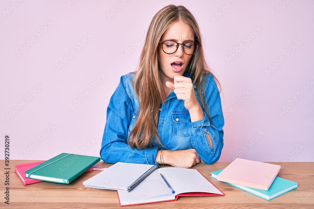 Young blonde girl studying sitting on the table feeling unwell and coughing as symptom for cold or bronchitis. health care concept. Young blonde girl studying sitting on the table feeling unwell and coughing as symptom for cold or bronchitis. health care concept.