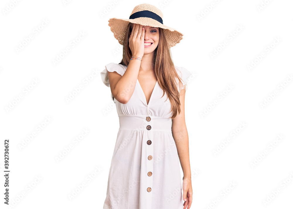 Young beautiful blonde woman wearing summer dress and hat covering one eye with hand, confident smile on face and surprise emotion. Young beautiful blonde woman wearing summer dress and hat covering one eye with hand, confident smile on face and surprise emotion.
