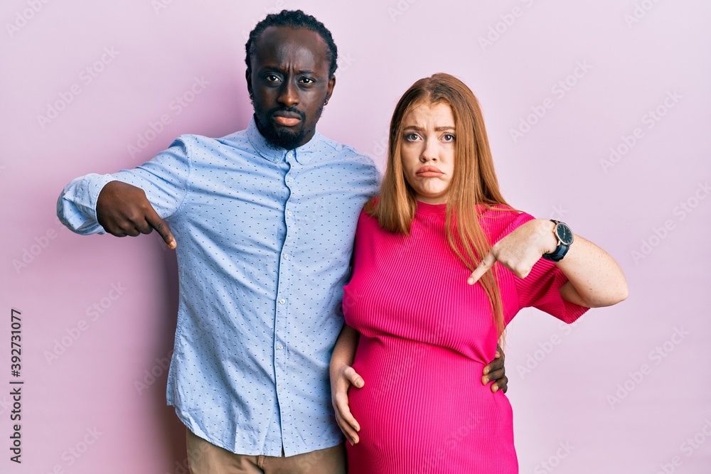 Obraz premium Young interracial couple expecting a baby, touching pregnant belly pointing down looking sad and upset, indicating direction with fingers, unhappy and depressed.