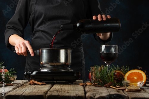 Close-up view of chef pours red wine into the pot for preparing mulled wine on rustic wooden table with festive composition background. Backstage of cooking hot drink with spices. Frozen motion.