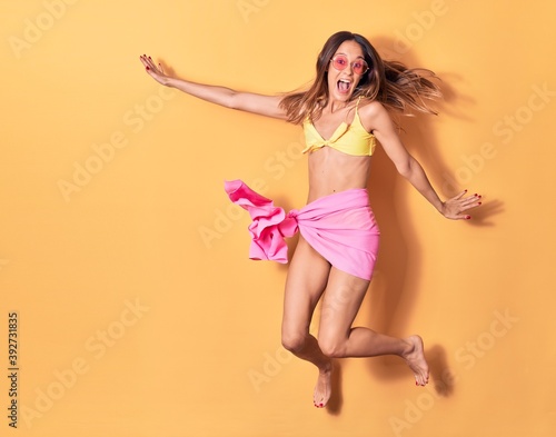 Young beautiful girl wearing bikini and sunglasses smiling happy. Jumping with smile on face over isolated yellow background