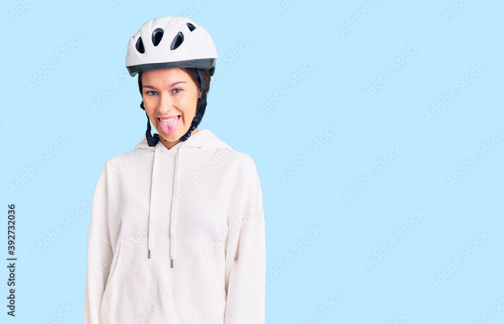 Beautiful brunette young woman wearing bike helmet and sporty clothes sticking tongue out happy with funny expression. emotion concept. Beautiful brunette young woman wearing bike helmet and sporty clothes sticking tongue out happy with funny expression. emotion concept.