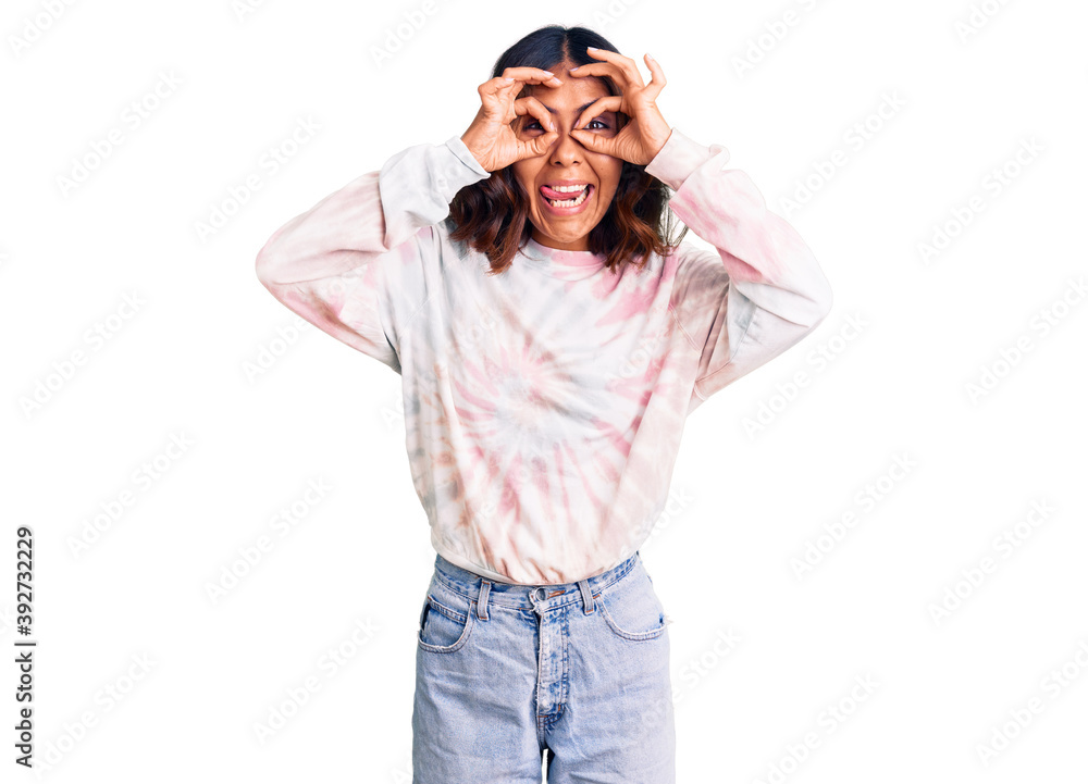 Young beautiful mixed race woman wearing casual tie dye sweatshirt doing ok gesture like binoculars sticking tongue out, eyes looking through fingers. crazy expression. Young beautiful mixed race woman wearing casual tie dye sweatshirt doing ok gesture like binoculars sticking tongue out, eyes looking through fingers. crazy expression.