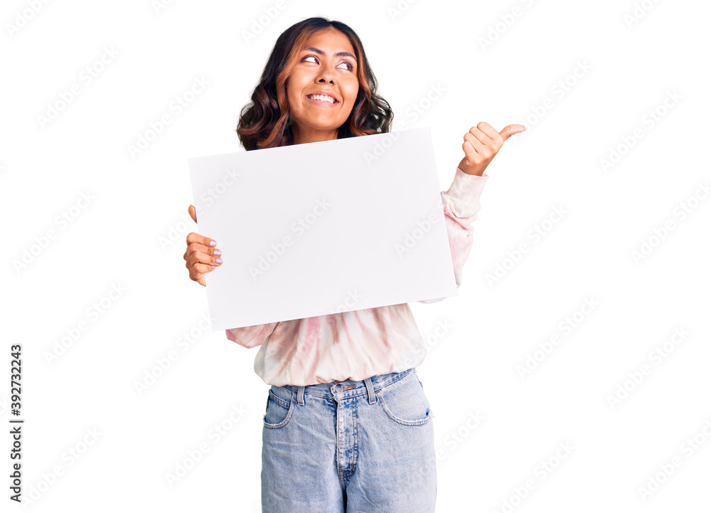 Young beautiful mixed race woman holding cardboard banner with blank space pointing thumb up to the side smiling happy with open mouth Young beautiful mixed race woman holding cardboard banner with blank space pointing thumb up to the side smiling happy with open mouth