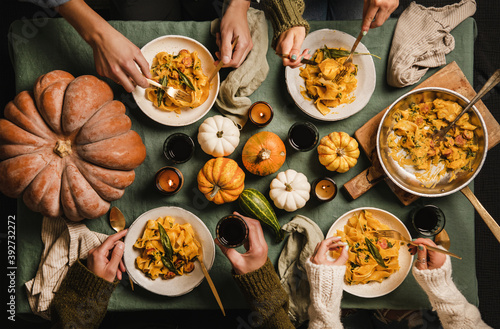 Flat-lay of Autumn dinner for gathering or Thanksgiving Day celebration. Family or friends eating butternut squash pasta with sausage and sage over tablecloth decorated with pumpkins, top view