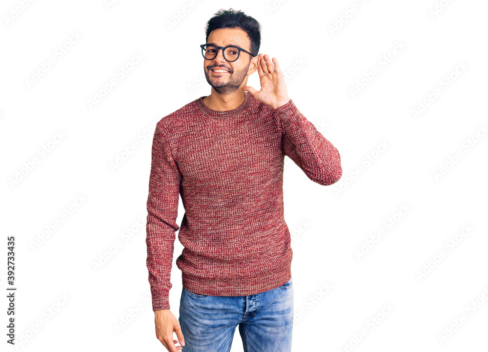Fototapeta premium Young handsome hispanic man wearing winter sweater and glasses smiling with hand over ear listening an hearing to rumor or gossip. deafness concept.
