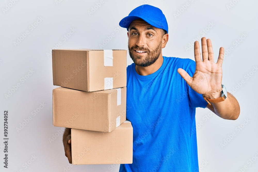 Handsome man with beard wearing courier uniform holding delivery packages waiving saying hello happy and smiling, friendly welcome gesture Handsome man with beard wearing courier uniform holding delivery packages waiving saying hello happy and smiling, friendly welcome gesture
