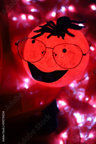 A very creepy cartoon moon shaped round light wearing glasses and a tarantula shaped spider over it's head smiling isolated on red background © FCEalin 