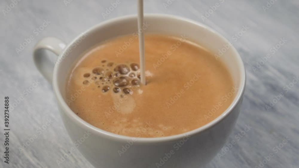 Pouring milk into coffee with foam in a white cup on a gray wooden table. Slow motion. Close-up Pouring milk into coffee with foam in a white cup on a gray wooden table. Slow motion. Close-up