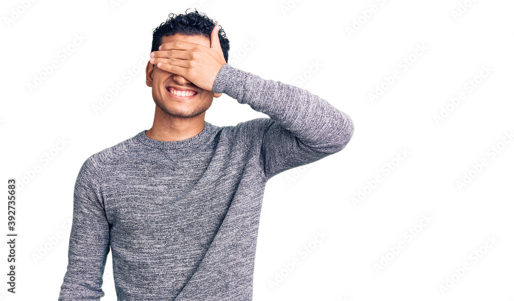 Hispanic handsome young man wearing casual sweater smiling and laughing with hand on face covering eyes for surprise. blind concept. Hispanic handsome young man wearing casual sweater smiling and laughing with hand on face covering eyes for surprise. blind concept.