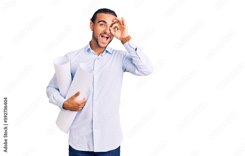 Young handsome man holding paper blueprints smiling happy doing ok sign with hand on eye looking through fingers Young handsome man holding paper blueprints smiling happy doing ok sign with hand on eye looking through fingers