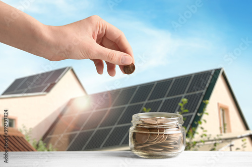 Woman putting coin into jar against house with installed solar panels on roof, closeup. Economic benefits of renewable energy photo
