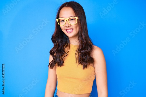 Young beautiful chinese girl wearing casual clothes and glasses looking positive and happy standing and smiling with a confident smile showing teeth