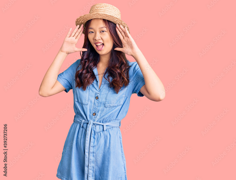 Young beautiful chinese girl wearing summer hat smiling cheerful playing peek a boo with hands showing face. surprised and exited Young beautiful chinese girl wearing summer hat smiling cheerful playing peek a boo with hands showing face. surprised and exited