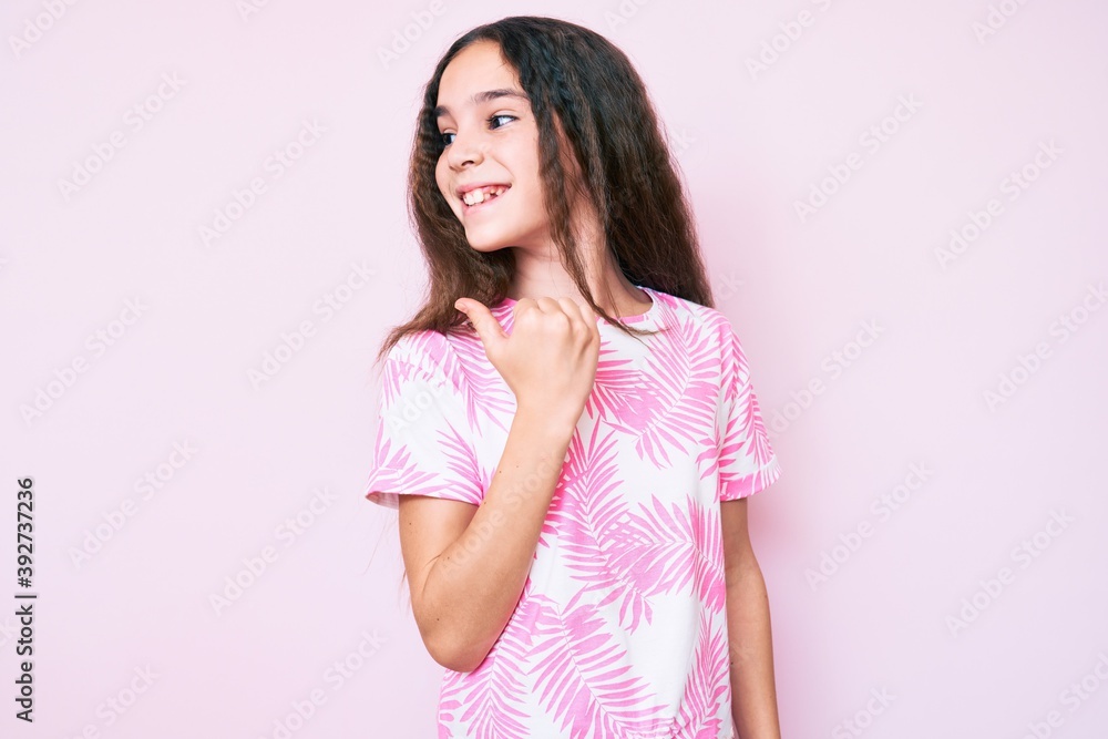 Cute hispanic child girl wearing casual clothes smiling with happy face looking and pointing to the side with thumb up. Cute hispanic child girl wearing casual clothes smiling with happy face looking and pointing to the side with thumb up.