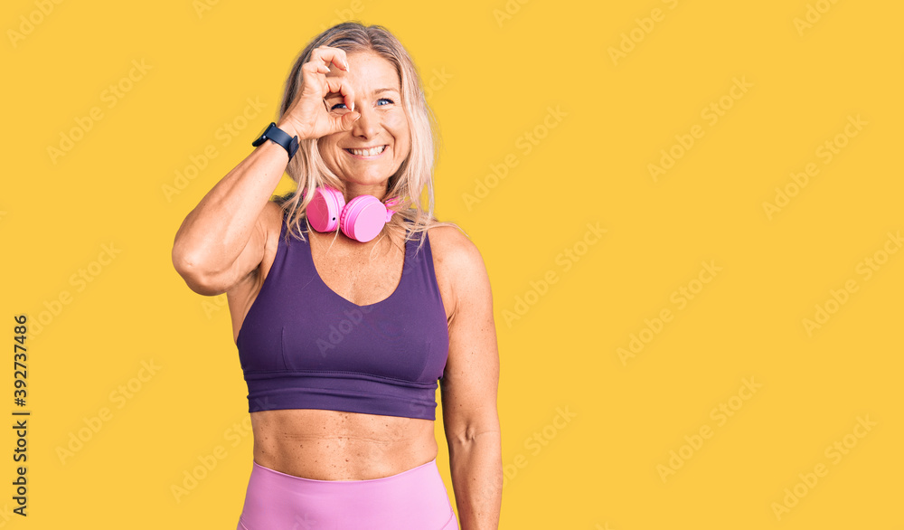 Middle age fit blonde woman wearing gym clothes and using headphones doing ok gesture with hand smiling, eye looking through fingers with happy face. Middle age fit blonde woman wearing gym clothes and using headphones doing ok gesture with hand smiling, eye looking through fingers with happy face.