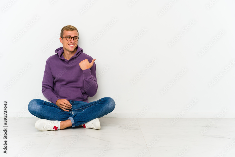 Young caucasian man sitting on the floor points with thumb finger away, laughing and carefree. Young caucasian man sitting on the floor points with thumb finger away, laughing and carefree.