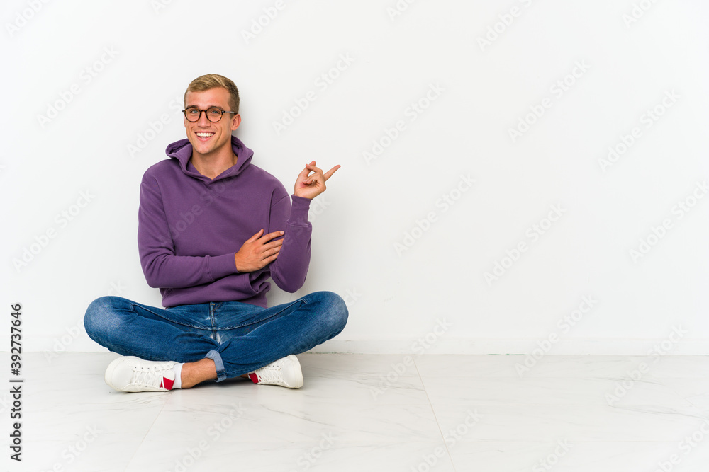 Young caucasian man sitting on the floor smiling cheerfully pointing with forefinger away. Young caucasian man sitting on the floor smiling cheerfully pointing with forefinger away.